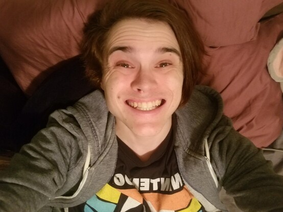 picture right before rye cut their hair and started parting down the middle -- swiped to the right. they are smiling, full-teeth, lying down and looking up at the camera wearing a blue sweater and an n64 shirt